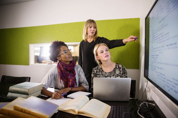 Tre kvinnliga forskare i kontorsmiljö. En äldre forskare står och pekar på en tavla på väggen med text, medn två mer juniora forskare som sitter vid datorn tittar på tavlan och lyssnar på den äldre forskaren som säger något.