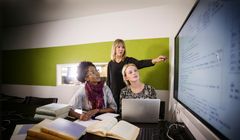 Tre kvinnliga forskare i kontorsmiljö. En äldre forskare står och pekar på en tavla på väggen med text, medn två mer juniora forskare som sitter vid datorn tittar på tavlan och lyssnar på den äldre forskaren som säger något.