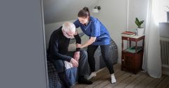 Kvinnlig hemtjänstpersonal hemma hos äldre man hjälper den äldre mannen att resa sig upp från sängen.