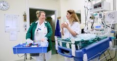Två sjukvårdspersonal, en i gröna kläder och en i vita, pratar med varandra i en sjukhuskorridor bredvid en medicinvagn och medicinsk utrustning.