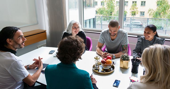 En grupp med fem personer som ser glada ut sitter runt ett bord i ett ljust konferensrum och samtalar med varandra.