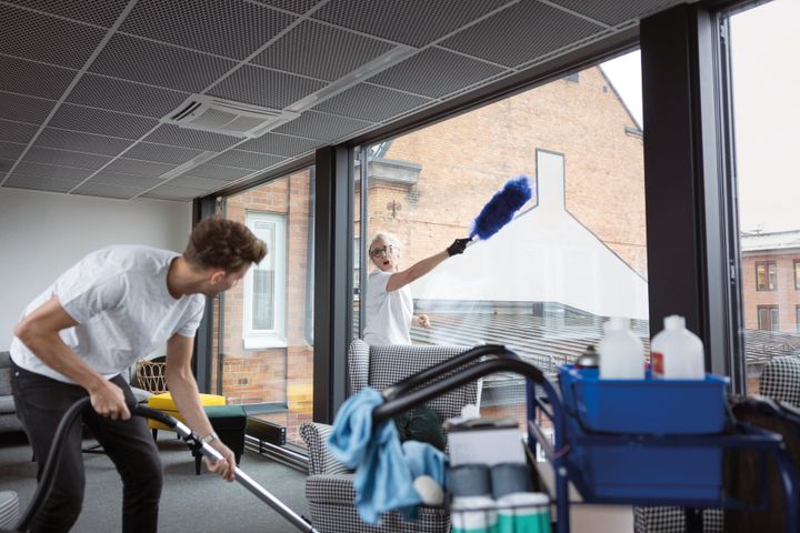 Två personer utför städning i ett kontor med stora fönster. En av dem dammsuger medan den andra tvättar fönster.