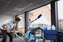 Två personer utför städning i ett kontor med stora fönster. En av dem dammsuger medan den andra tvättar fönster.
