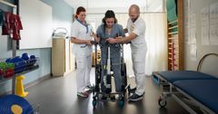 En person får hjälp att gå med en rullator av två vårdpersonal på en ortopedavdelning på ett sjukhus.