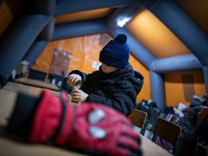 Ett barn klädd i vinterkläder häller något från en termos i ett uppvärmt tält. En röd handske ligger på bordet i förgrunden.