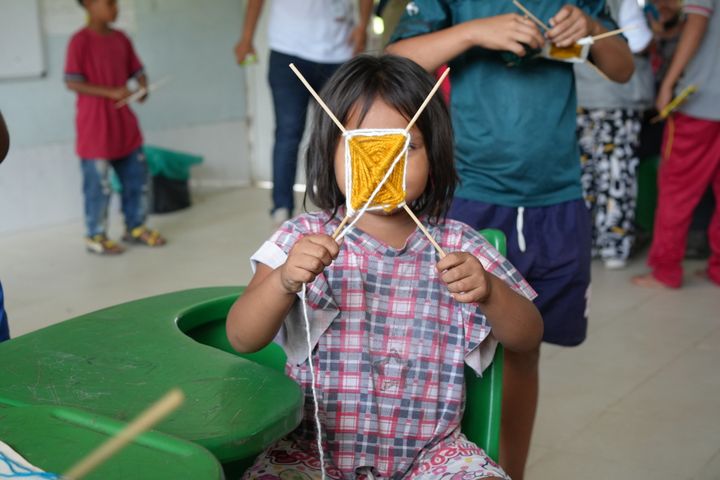 En ung flicka visar stolt upp sin konstverk i ett klassrum. Hon och andra barn deltar i en aktivitet där de skapar med garn och pinnar.
