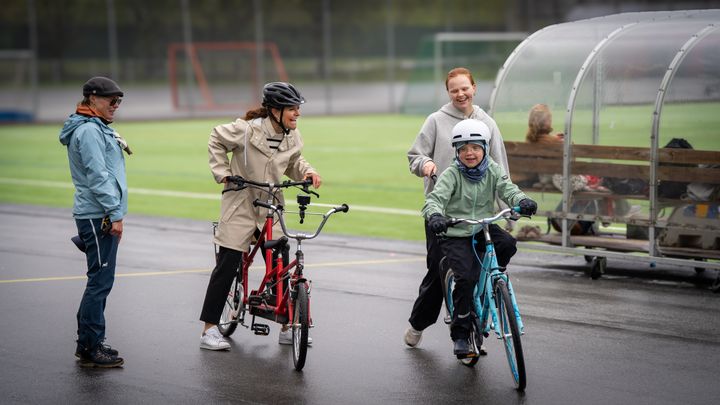 En grupp människor cyklar tillsammans på en idrottsplats. En person hjälper en yngre cyklist. Alla bär hjälmar.