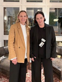 Två kvinnor står tillsammans inomhus, den ena i beige kavaj och den andra i svart kavaj.