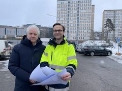 Två män iklädda vinterjackor står utomhus vid Mörbyplan med skisser i handen, med höghus och bilar i bakgrunden.