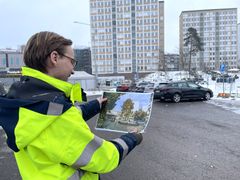 En person i gula arbetskläder håller upp en bild av ett framtida byggprojekt framför ett parkeringsområde med höghus i bakgrunden.