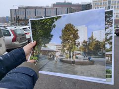 En hand håller en bild som visar en framtida vision av Mörbyplan, med moderna byggnader och grönska, medan den nuvarande platsen syns i bakgrunden.