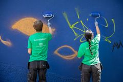 Två ferieungdomar står med ryggen till och målar över grafitti på en blå vägg. De har på sig svarta arbetarbyxor och gröna t-shirts med texten Sommarjobbare och Familjebostäders logga.