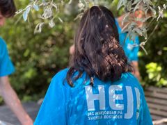 En person med långt svart hår och en blå t-shirt med texten "Hej! Jag feriejobbar på Svenska Bostäder" fotad bakifrån