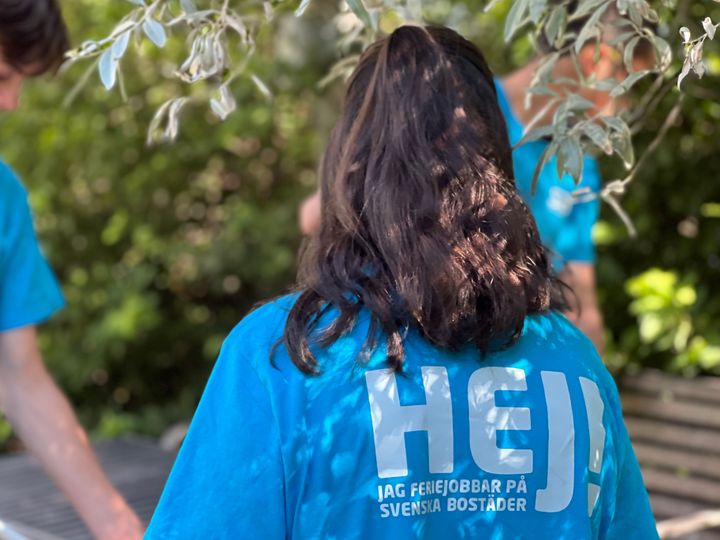 En person med långt svart hår och en blå t-shirt med texten "Hej! Jag feriejobbar på Svenska Bostäder" fotad bakifrån