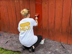 En person med blont hår uppsatt i knut, iförd vit t-shirt med texten "Hej! Jag feriejobbar på Stadsholmen" målar ett rött staket