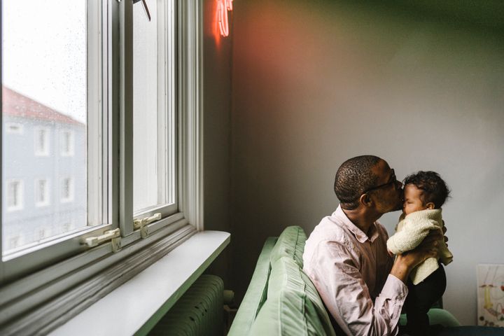Bilden visar en man som lyfter upp en bebis i famnen, sittandes i en grön soffa som står mot fönstret i en skralt inredd lägenhet.