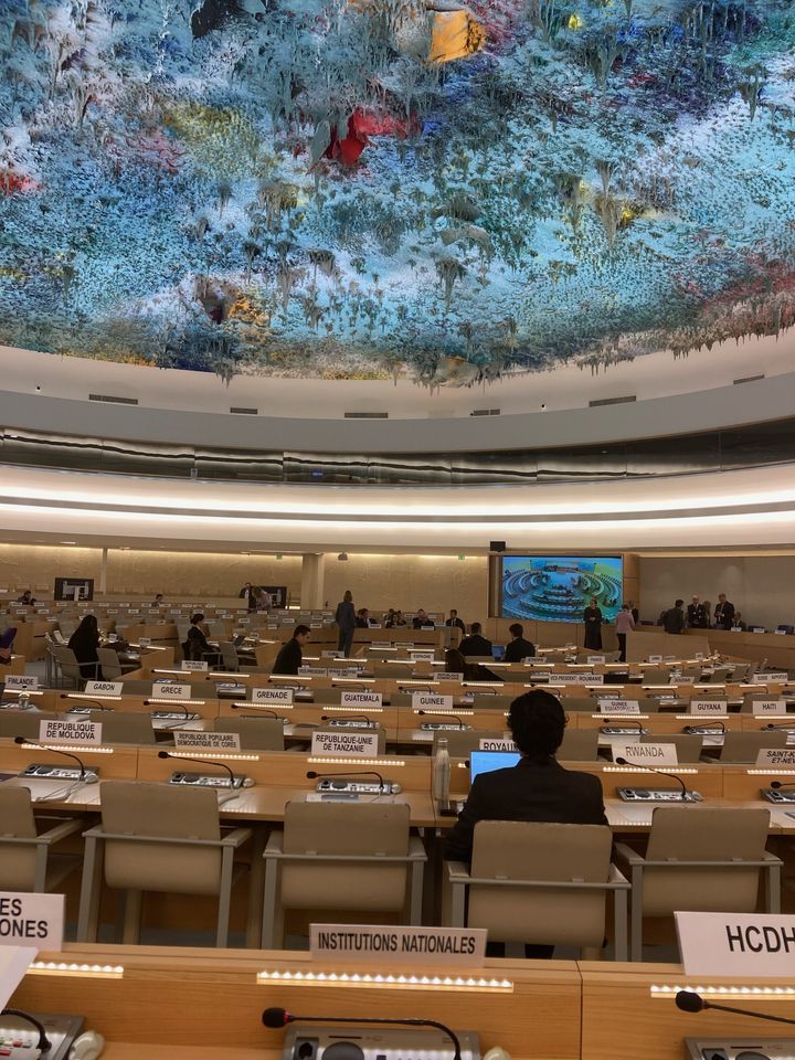 Konferensrum på FN:s kontor i Genève. Rader med stolar. Människor står och sitter i rummet. I rummet finns också en takskulptur av den spanska konstnären Miquel Barceló. Stalaktitformade droppar hänger från skulpturen i blåa nyanser.