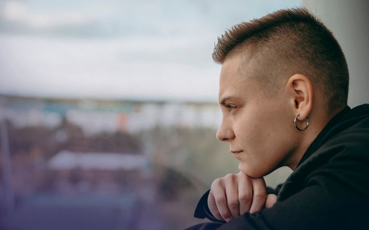 Ung person blickar ut genom ett fönster med utsikt över ett bostadsområde. Personen ser både betänksam och besluten ut. (Fotografi).