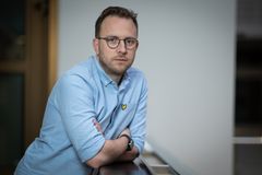 En man med glasögon, i en ljusblå skjorta, står lutad mot ett räcke inomhus.