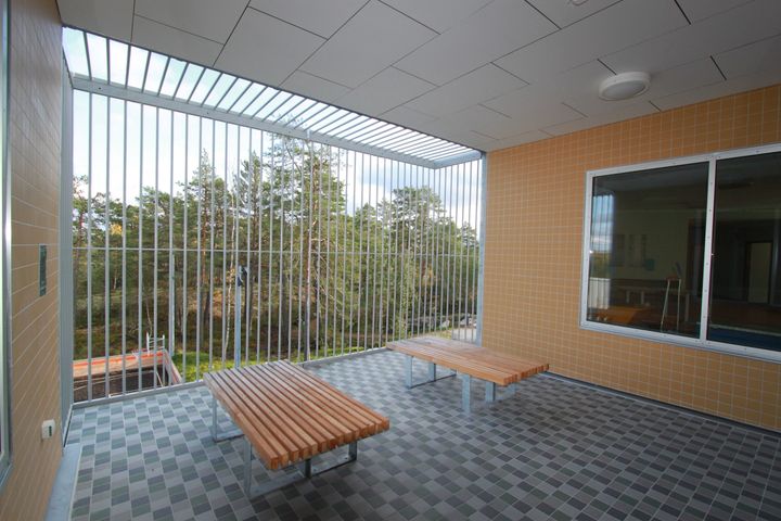 Alt text: Utrymme med träbänkar och rutigt golv i rättspsykiatrisk enhet, utsikt över träd.