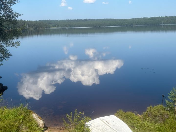 En klar sjö med spegelbild av moln på ytan, omgiven av skog.