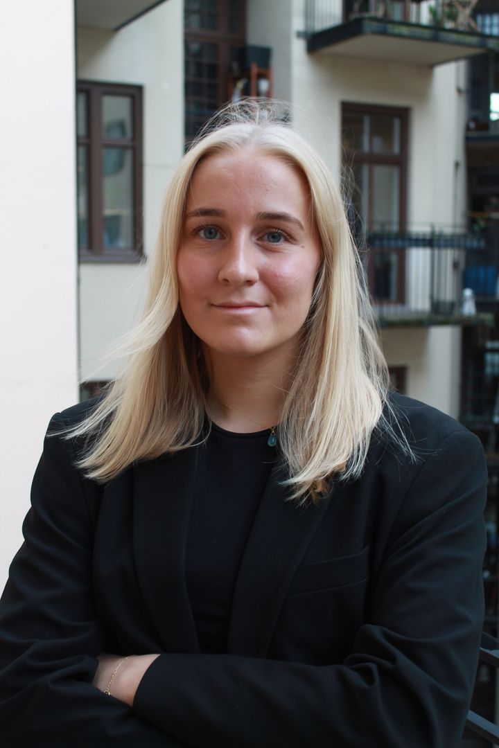 Kvinna%20med%20l%E5ngt%20blont%20h%E5r%2C%20svart%20kavaj%2C%20st%E5r%20framf%F6r%20en%20byggnadsinteri%F6r.