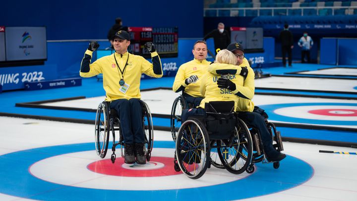 Fyra personer med gula landslagströjor i varsin rullstol på en curlingbana där tre personer kramas och en sticker armarna uppåt i luften.