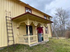 En äldre man i röd tröja står på verandan till ett gult trähus på landet.