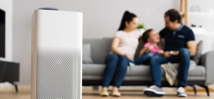 En vit luftrenare i förgrunden, med en familj som sitter på en soffa i bakgrunden.
