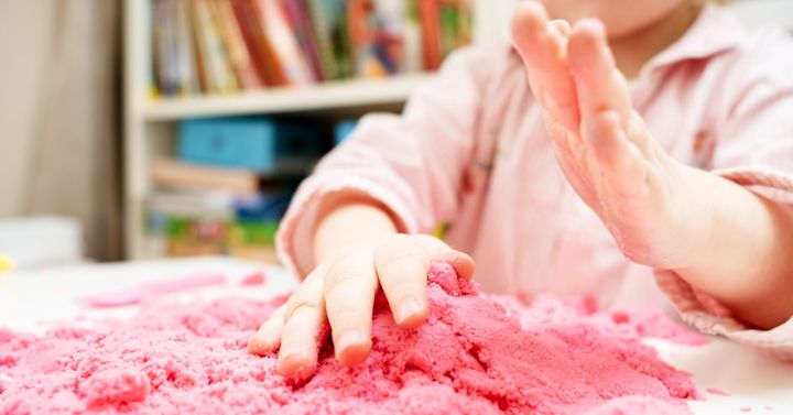 På bilden syns ett barn som leker med rosa leksand. Det är en närbild där mest armar och händer syns.