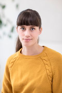 En portätt bild på Frida Ramström som har mörkt hår i hästsvans och en senapsgul stickad tröja på sig.