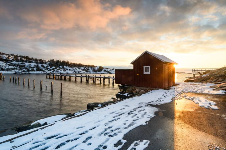 Litet rött fritidshus vid vattnet och en brygga med snötäckt mark.