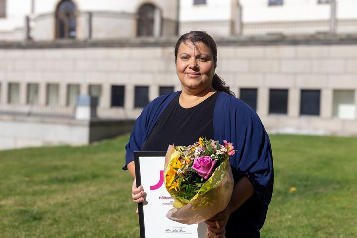 Kvinna håller blombukett och diplom, står utomhus framför en byggnad.