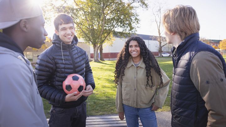 Fyra ungdomar står utomhus i en park och pratar. En av dem håller en röd fotboll.
