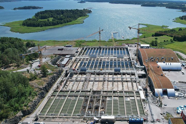 Flygbild av ett reningsverk vid vattnet, med flera byggnader och kranar i området.