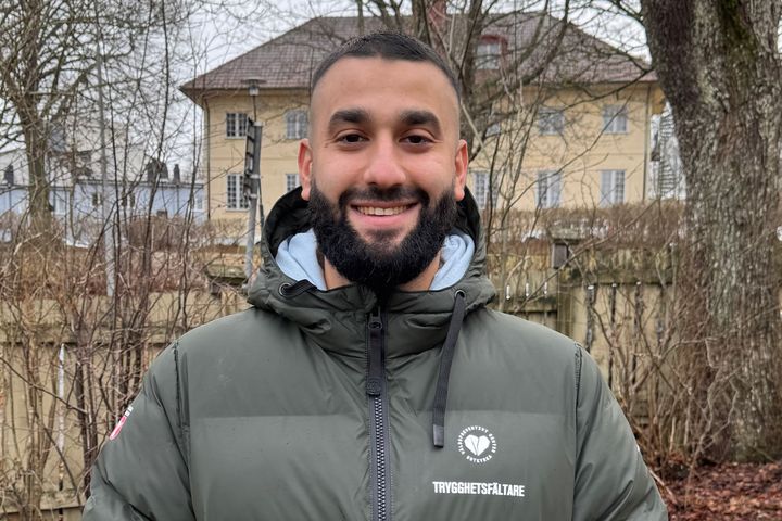 En person står utomhus i en grön jacka med texten "Trygghetsvärd" på bröstet. Bakom syns ett gult hus och träd utan löv.