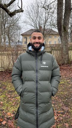 En person står utomhus i en park klädd i en lång, grön vinterjacka. Bakgrunden består av träd och ett hus.