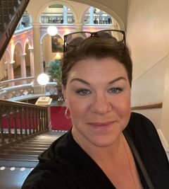 En kvinna i glasögon som står inomhus framför en trappa med en dekorativ inredning i bakgrunden.