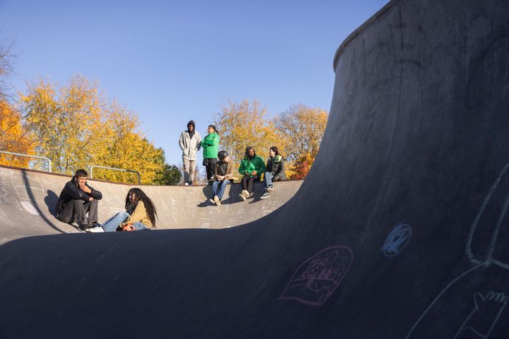 En grupp människor sitter och står i en skateboardramp på en solig dag, med träd i höstfärger i bakgrunden.