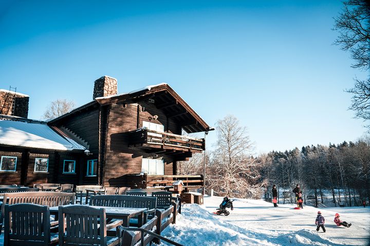 En snöig vinterdag vid en trästuga, med barn som leker på en pulkabacke i närheten.