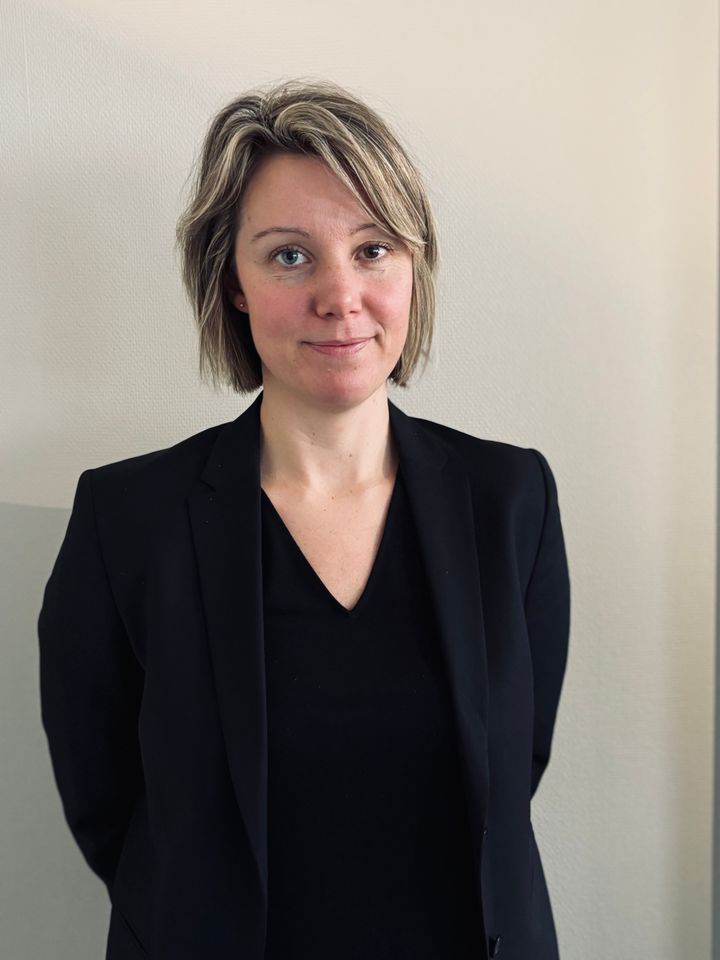 Kvinna med ljust hår i svart kavaj mot en ljus vägg.