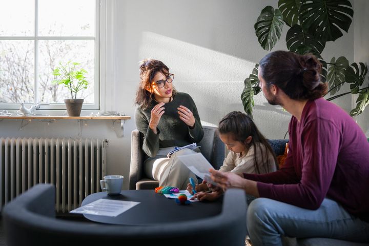 Tre personer sitter och pratar i ett vardagsrum. En vuxen kvinna gestikulerar medan en man håller ett papper. Ett barn ritar vid bordet.