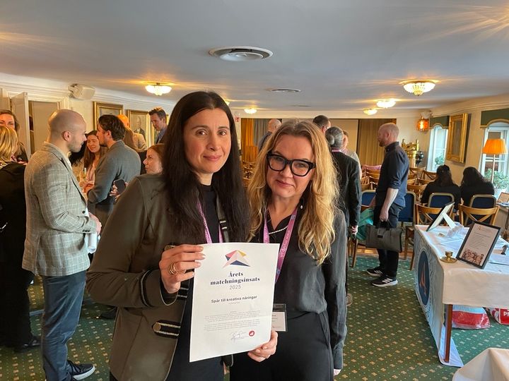 Två personer står i ett rum och håller ett diplom. Det finns fler personer och bord i bakgrunden.