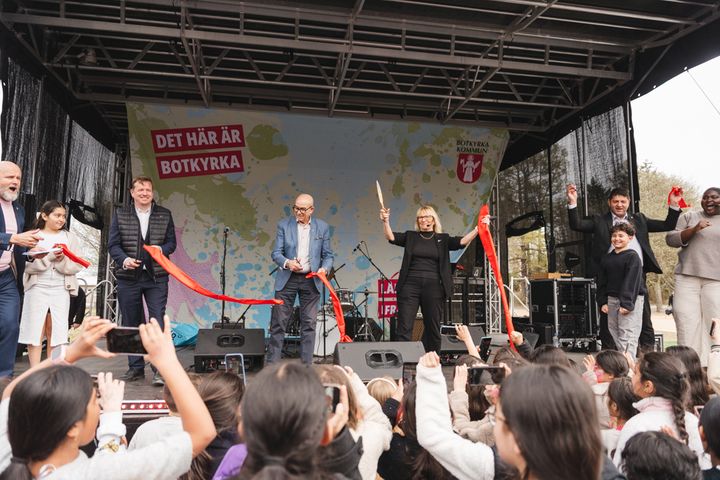 En grupp personer klipper ett rött band på en utomhusscen, omgivna av en glad publik.
