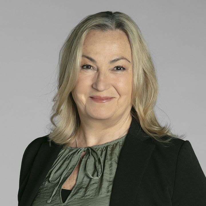 En person med blont hår ler mot kameran. De har en grön blus och svart kavaj.