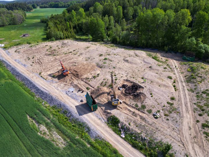Flygbild av grävmaskiner som arbetar på en sandig byggarbetsplats intill gröna fält och skog.