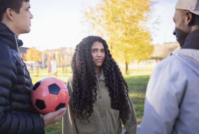 Tre ungdomar står på en fotbollsplan, en håller en fotboll.