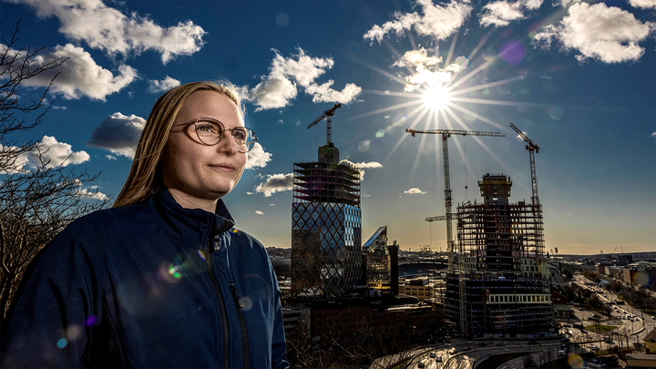 En kvinna står i förgrunden till vänster i bilden, i bakgrunden lyser solen över en byggnad med glasfasad som är under uppbyggnad