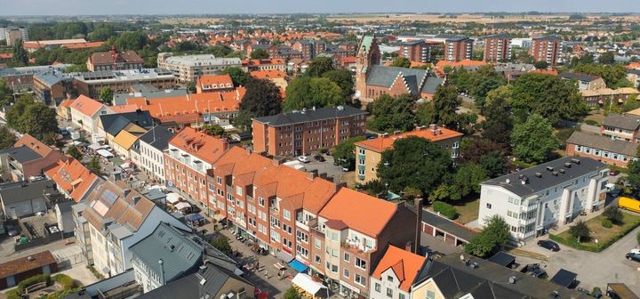 Flygbild av en stad med tegel- och vita hus, omgiven av grönska och flerfamiljshus i bakgrunden.