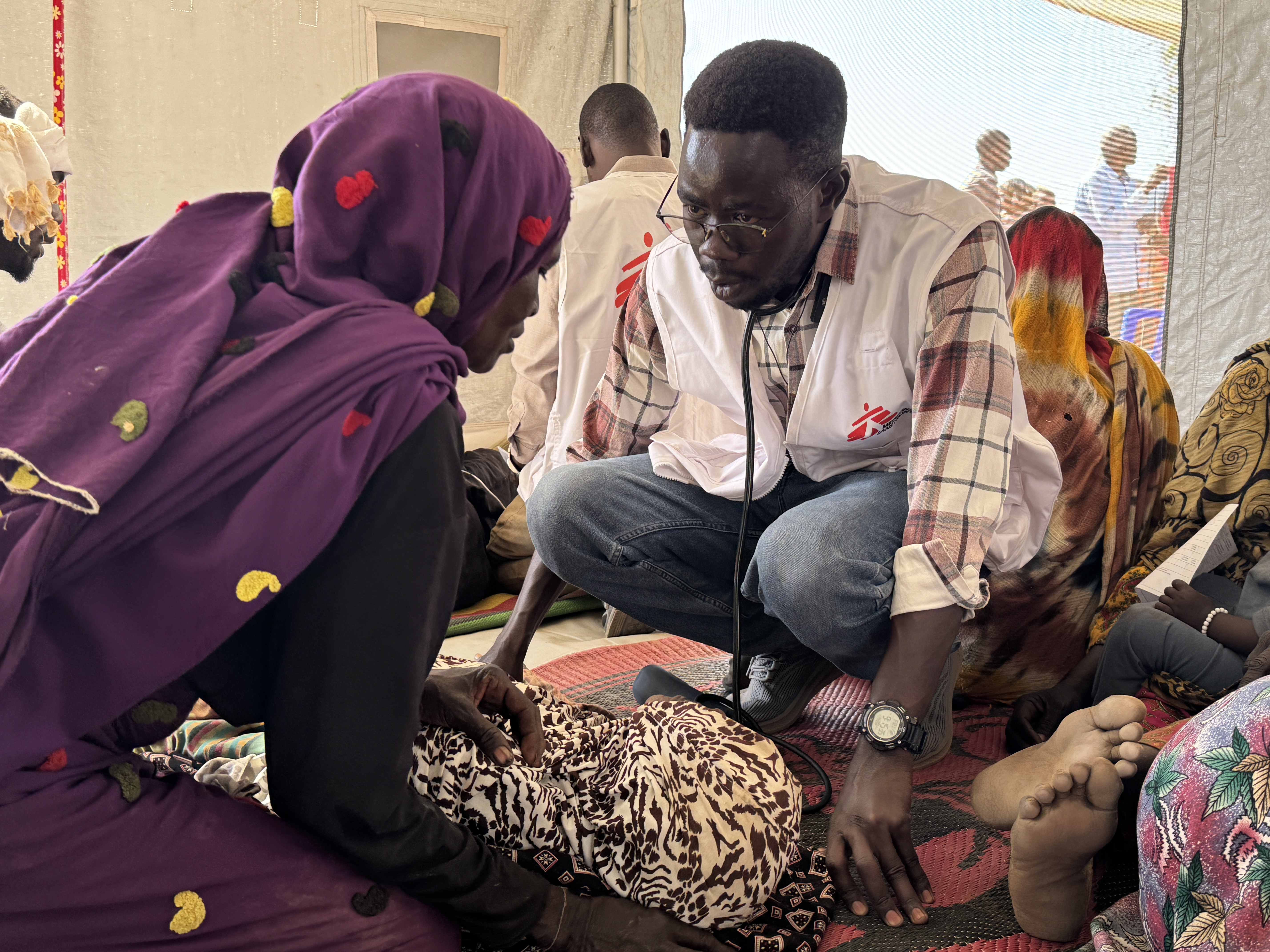Läkaren Abdel Mafid sitter på huk vid en äldre kvinna på Läkare Utan Gränsers enklare vårdcentral i Tawila.